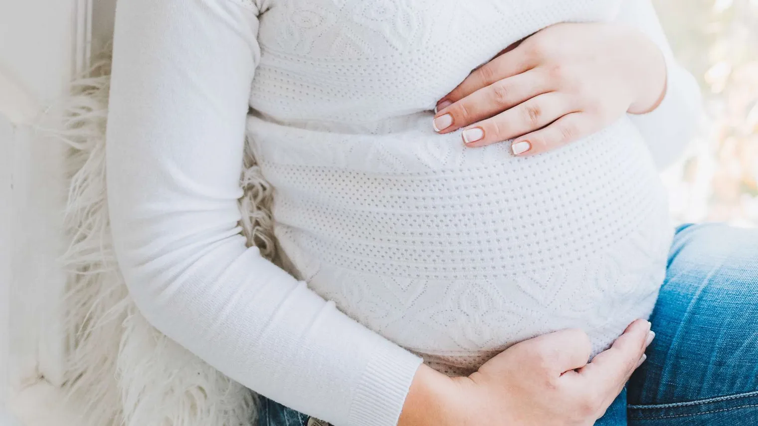 Schwangere Frau in weißem Strickpullover und Jeans sitzt auf einem hellen Fell und hält mit beiden Händen ihren Bauch