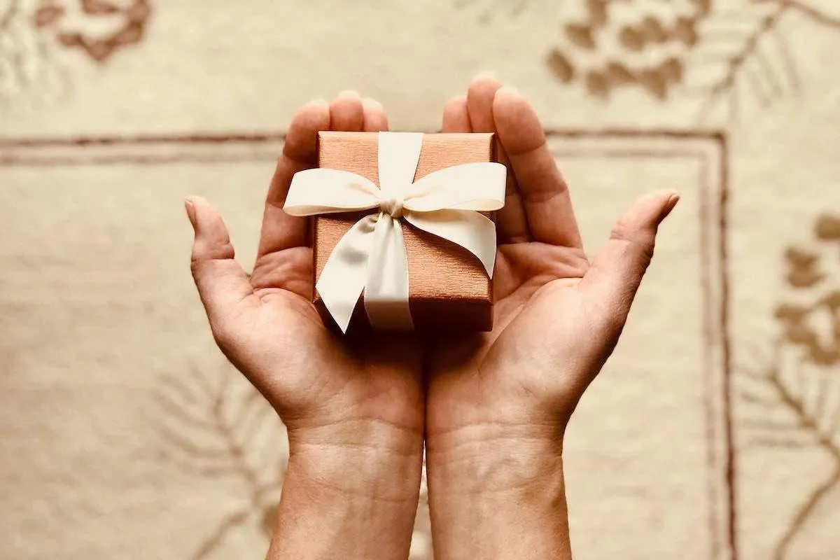 Zwei geöffnete Hände halten ein kleines kupferfarbenes Geschenk mit cremefarbener Schleife über einem gemusterten Teppich