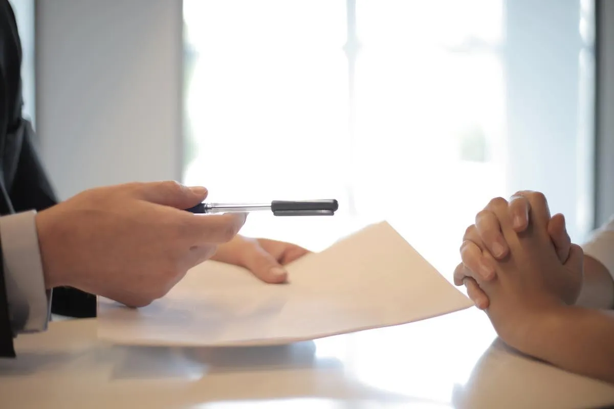 Hand im dunklen Anzug reicht einen Stift und ein Dokument über den Tisch zu einer Person mit gefalteten Händen