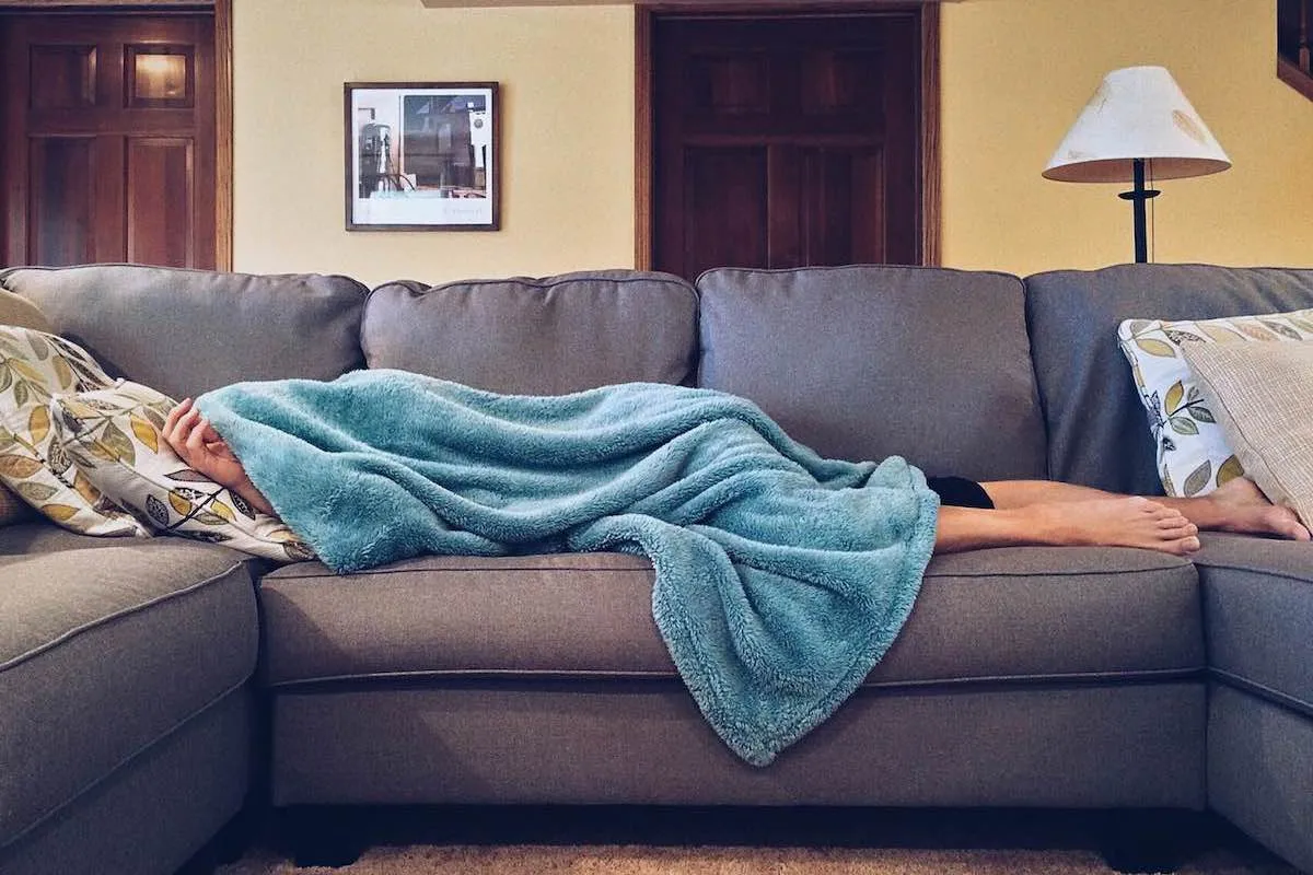 Eine Person liegt unter einer türkisfarbenen Fleecedecke auf einem grauen Sofa, nur ein Arm und die nackten Füße ragen heraus